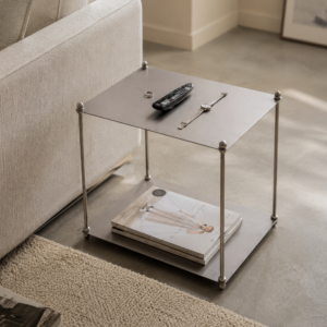 Small gunmetal side table with silver ball-joint legs beside a beige sofa, topped with a remote control, watch, and ring, with magazines stored on the lower shelf